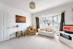 A living room with a white sofa, a wooden chair, a small table with a lamp, a painting on the wall, and a TV displaying 'GUEST READY'. - GuestReady - Modern Retreat with Classic Charm (London)