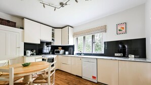 Moderni keittiö, jossa on valkoiset kaapit, puinen ruokapöytä ja ikkuna, josta avautuu näkymä vehreään maisemaan.