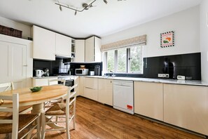 A modern kitchen with white cabinets, a wooden dining table, and a window with a view of greenery. - GuestReady - Modern Retreat with Classic Charm (London)