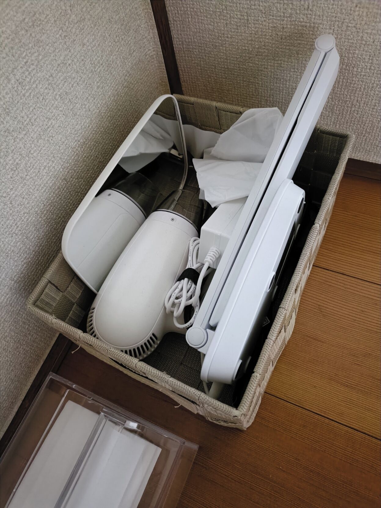 A wicker basket containing a hairdryer, a vacuum cleaner, and a white appliance, possibly a humidifier or air purifier, on a wooden floor.