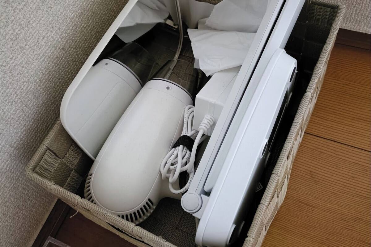 A wicker basket containing a hairdryer, a vacuum cleaner, and a white appliance, possibly a humidifier or air purifier, on a wooden floor.