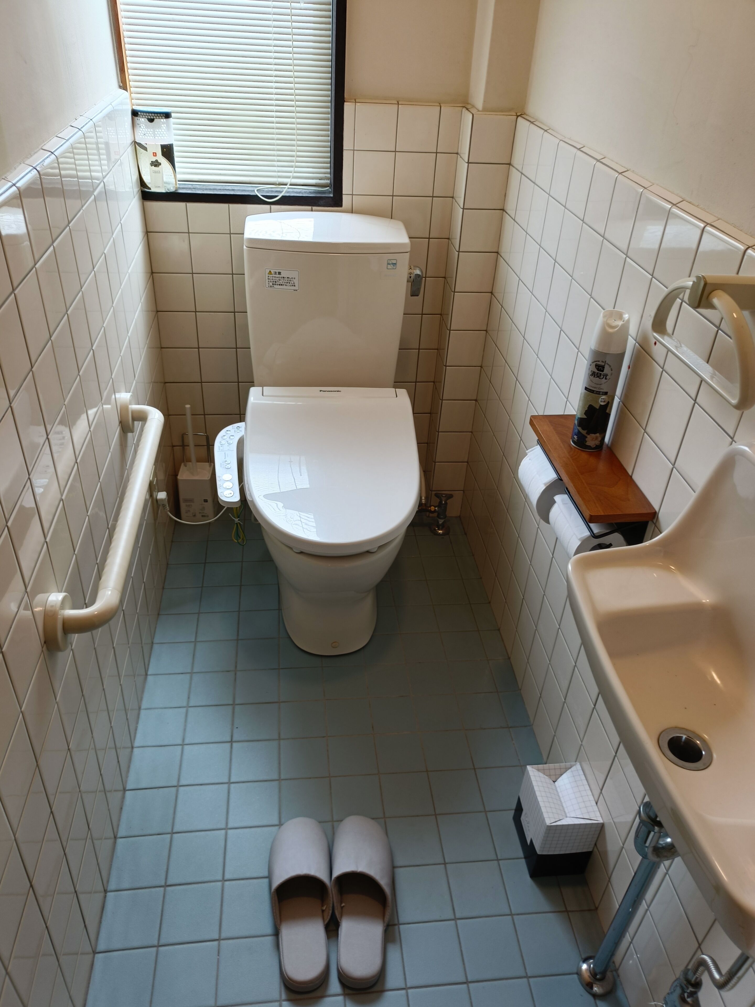 A small bathroom with a toilet, a handrail, a window, a shelf with toiletries, and a pair of slippers on the floor.