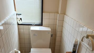 A small bathroom with a toilet, a handrail, a window, a shelf with toiletries, and a pair of slippers on the floor.