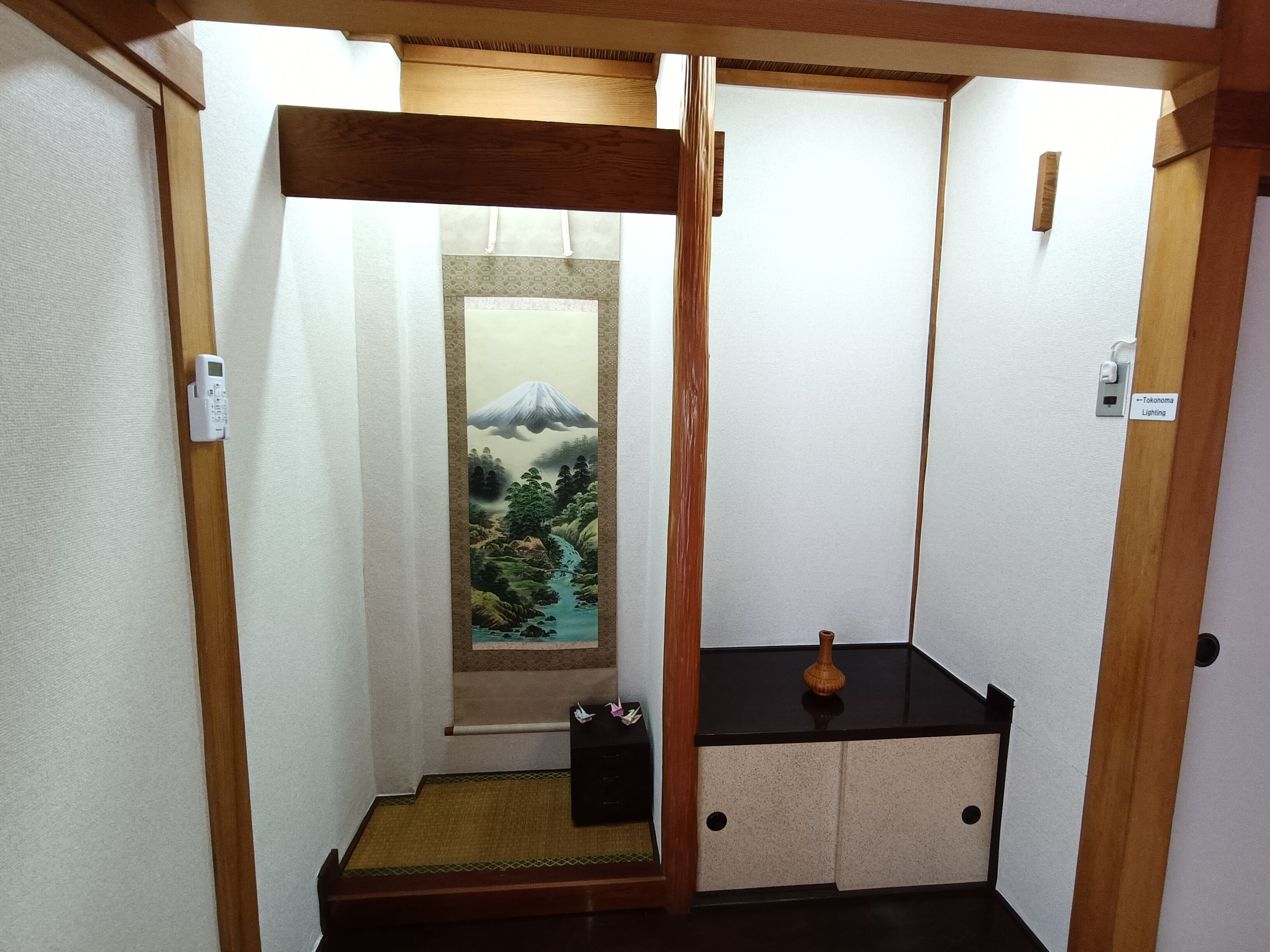 Un couloir étroit avec une porte coulissante qui donne sur une pièce où l’on trouve une porte coulissante traditionnelle japonaise ornée d’un paysage de montagne, une petite armoire noire et un banc en bois.