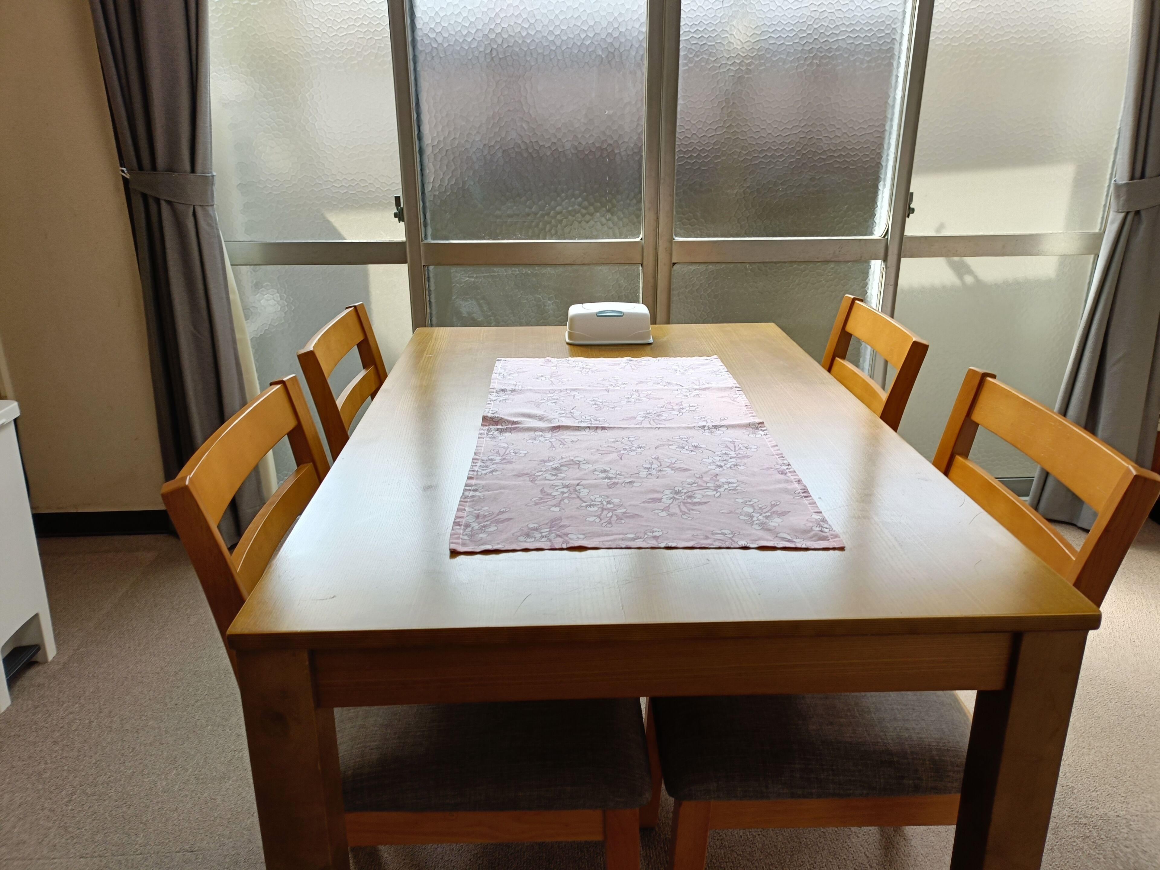 Una mesa de comedor de madera con camino de mesa floral, cuatro sillas de madera, un objeto blanco sobre la mesa y un ventanal de vidrio esmerilado al fondo.
