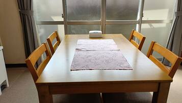 A wooden dining table with a floral table runner, four wooden chairs, a white object on the table, and a frosted glass window behind.