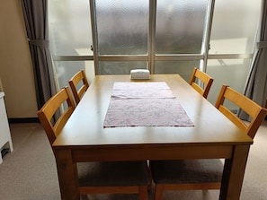 A wooden dining table with a floral table runner, four wooden chairs, a white object on the table, and a frosted glass window behind. - Shige no Sato Matsufuku (Takamatsu City)