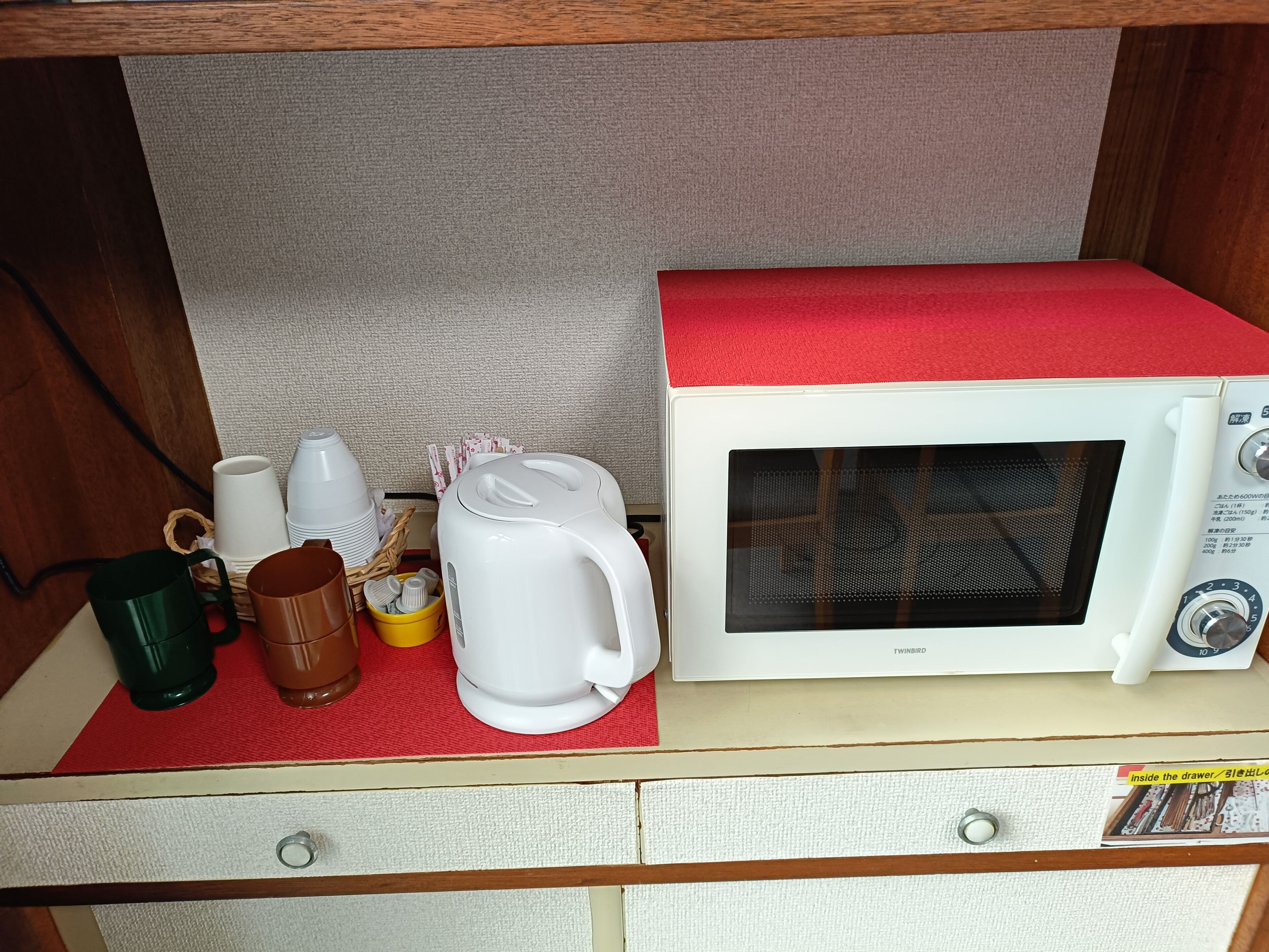 Un horno microondas con parte superior roja, una cafetera blanca, una taza verde y una taza amarilla sobre un tapete rojo dentro de un gabinete de madera.