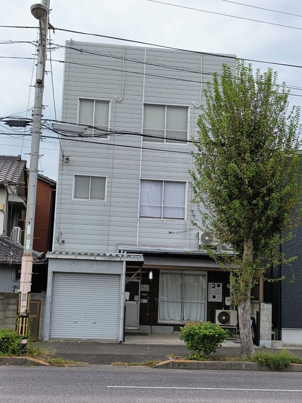 A two-story building with a garage door, air conditioning units, and a tree in front. - Shige no Sato Matsufuku (Takamatsu City)