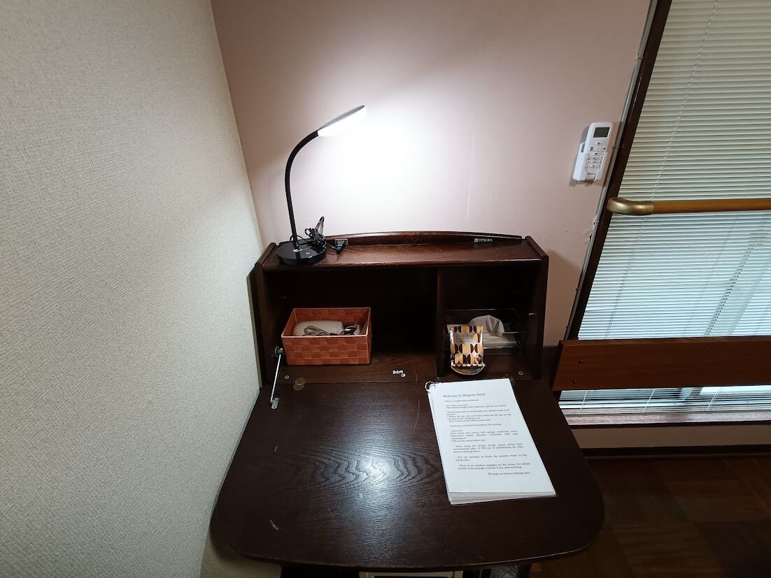 A small desk with a lamp, a notepad, and a window with blinds.