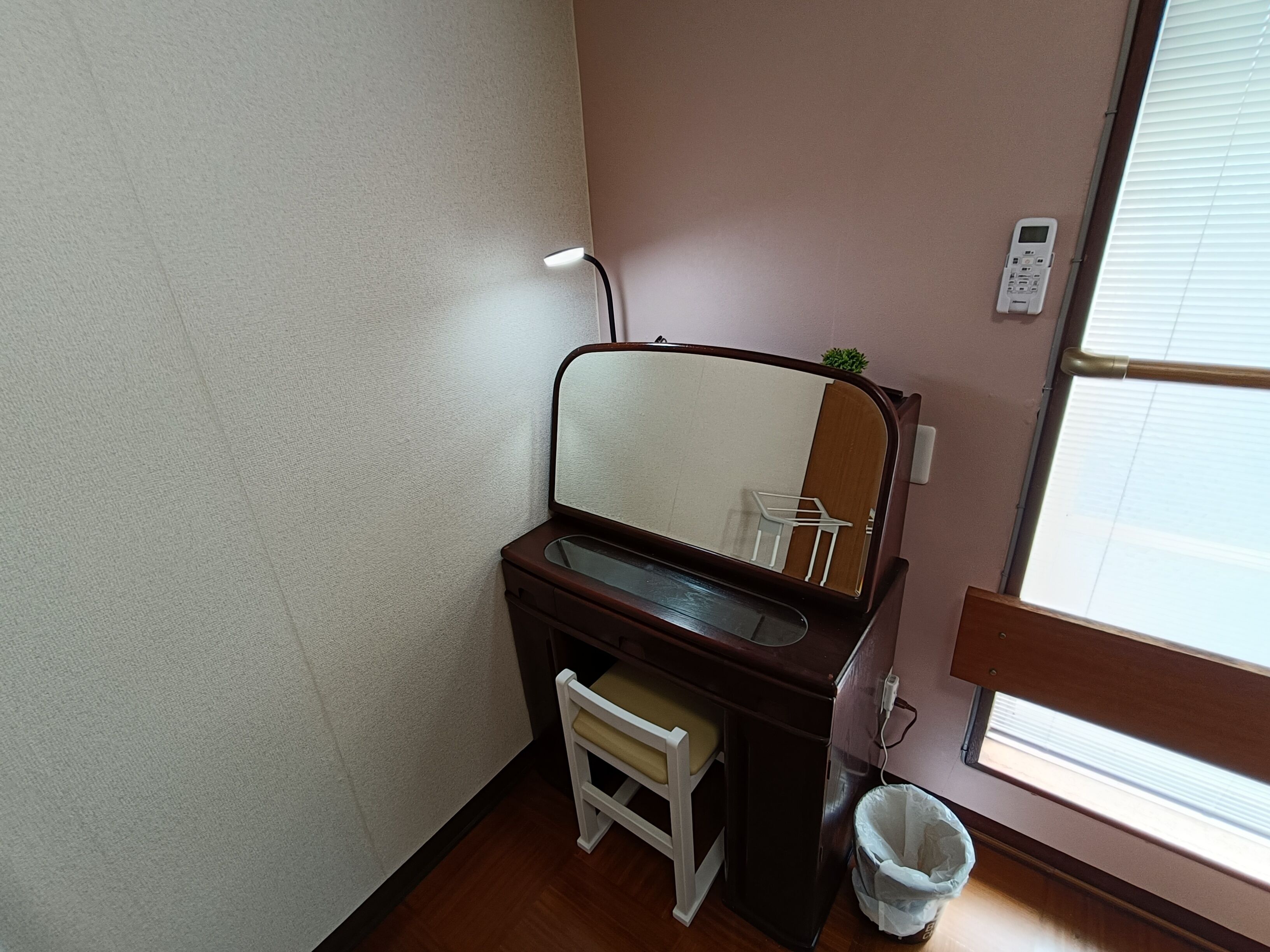 A bathroom with a wooden vanity, a mirror, a small stool, a trash bin, and a window with blinds.