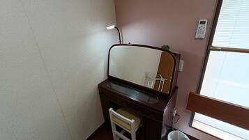 A bathroom with a wooden vanity, a mirror, a small stool, a trash bin, and a window with blinds.
