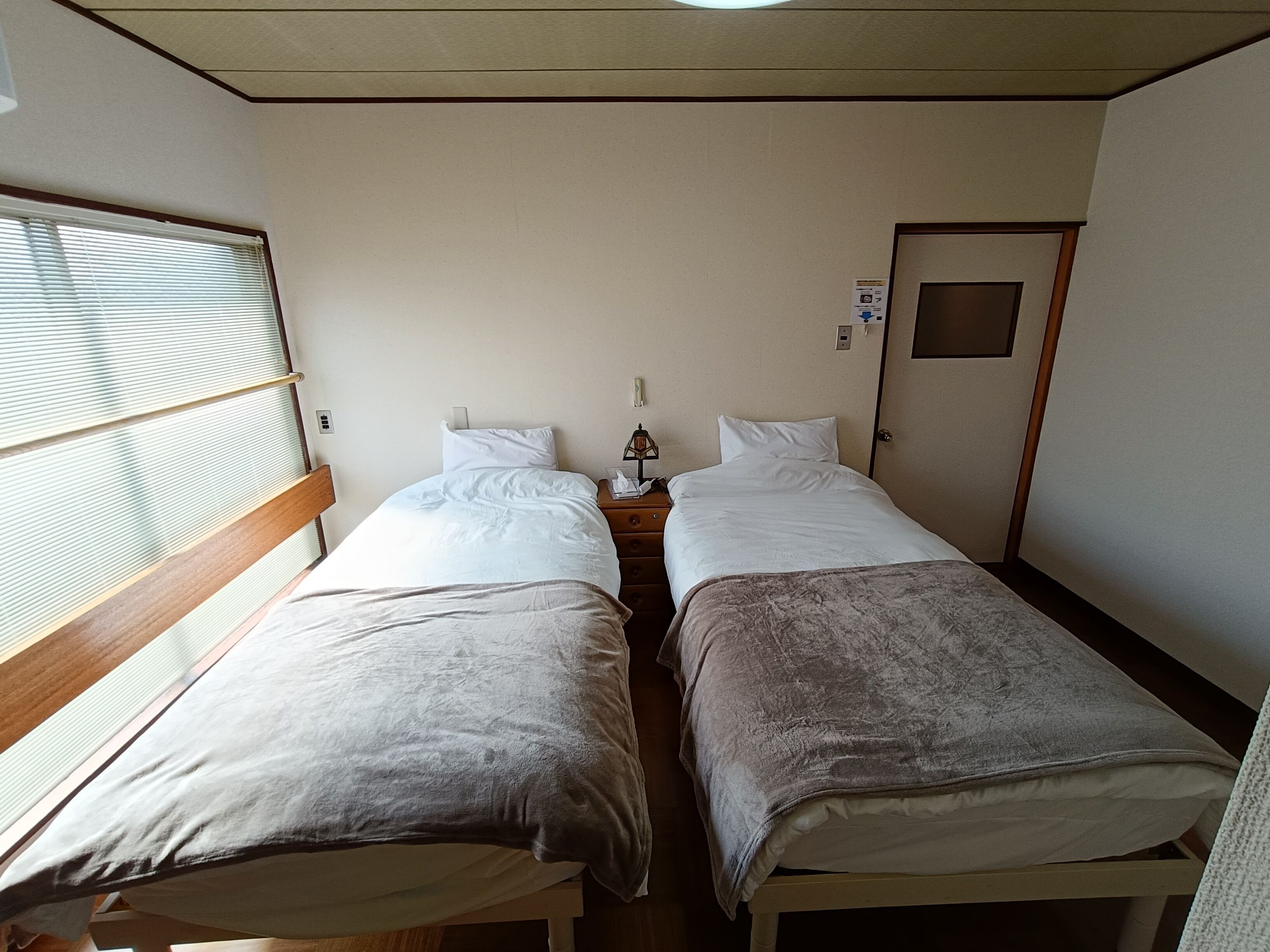 A room with two beds, a window with blinds, a door, and a small table with a lamp.