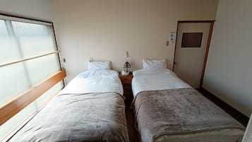 A room with two beds, a window with blinds, a door, and a small table with a lamp.