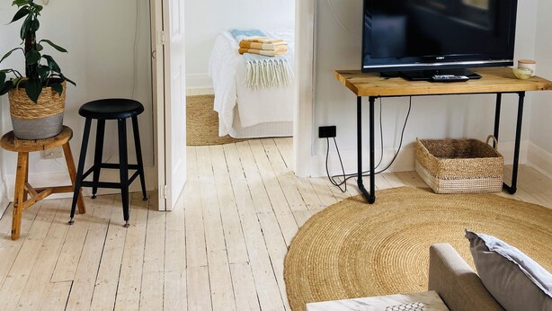 Uma sala de estar com televisão, mesa de madeira, sofá, planta em vaso e uma cama ao fundo.