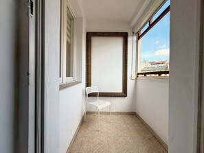 Ein schmaler Balkon mit einem weißen Stuhl, einem großen Spiegel mit Rahmen und Blick auf Gebäude durch ein Fenster.