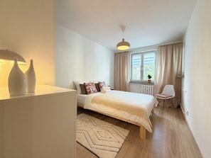 Ein Schlafzimmer mit einem Bett, einem Sessel, einem Heizkörper, einem Fenster mit Vorhängen und einer Pendelleuchte.