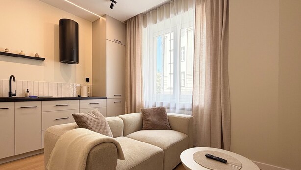 A modern living room with a sofa, a coffee table, a kitchen area, and a large window with curtains. - Stawowa Deluxe Apartment (Wrocław)