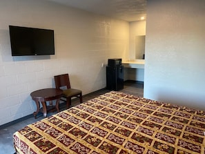Una habitación pequeña con una cama, una silla, un televisor en la pared y un refrigerador.