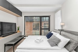 Ein modernes Hotelzimmer mit Bett, Fernseher, Schreibtisch und einem Balkon mit Blick auf Gebäude.