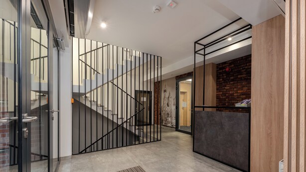 Un interno moderno con ingresso tramite porta in vetro, una scala con ringhiere nere e un'area reception caratterizzata da una parete in mattoni e pannelli in legno.