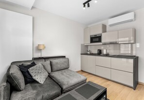 A modern living space with a grey sofa, a small table, a kitchen area with cabinets and appliances, and a wall-mounted air conditioner. - Main Station Comfort Studio (Kraków)