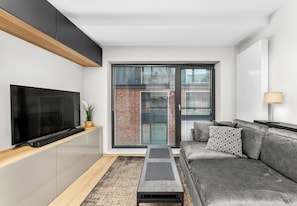 Ein modernes Wohnzimmer mit einem Flachbildfernseher, einem grauen Sofa, einem Couchtisch und einem großen Fenster mit Jalousien.