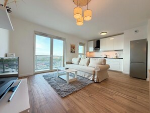 A modern apartment with a sofa, a coffee table, a kitchen area, and a large window with a city view. - Wrocław Partynice Cosy Apartment (Wrocław)