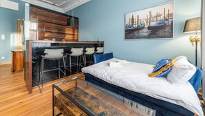 Una habitación con una cama, un escritorio, una silla, una zona de bar y un cuadro que representa una escena de canal.