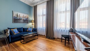 Una habitación con un sofá azul, una mesa de centro de madera, un escritorio y una silla. Hay un cuadro en la pared y una ventana con cortinas.