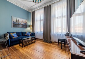 Ein Zimmer mit einem blauen Sofa, einem Holztisch, einem Schreibtisch und einem Stuhl. An der Wand hängt ein Gemälde und es gibt ein Fenster mit Vorhängen.