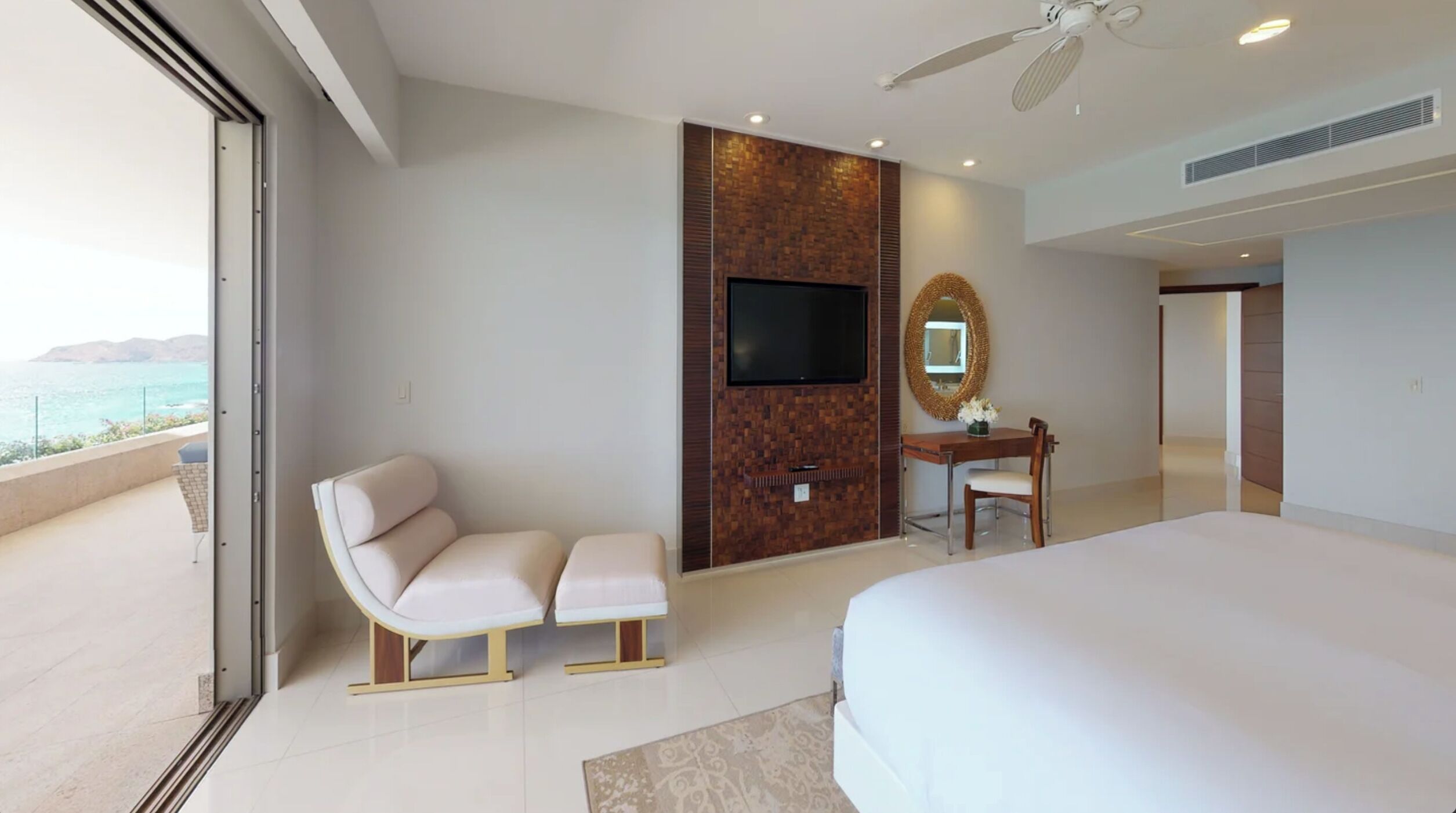 Ein modernes Hotelzimmer mit einem großen Bett, einer Sitzecke mit Sofa und Sesseln, einem Schreibtisch mit Stuhl, einem an einer Holzvertäfelung befestigten Flachbildfernseher und Blick aufs Meer.