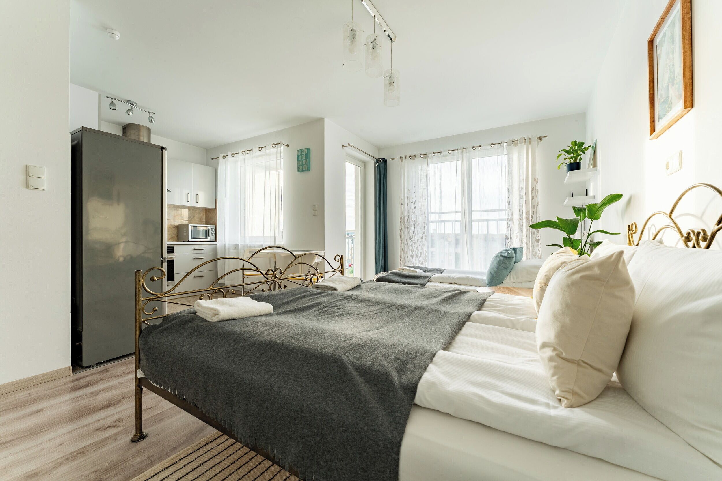 A bedroom with a bed, a bedside table, a window with curtains, a refrigerator, and a kitchen area.