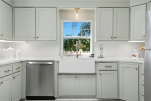 A kitchen with white cabinets, a stainless steel dishwasher, and a window with a view of trees.