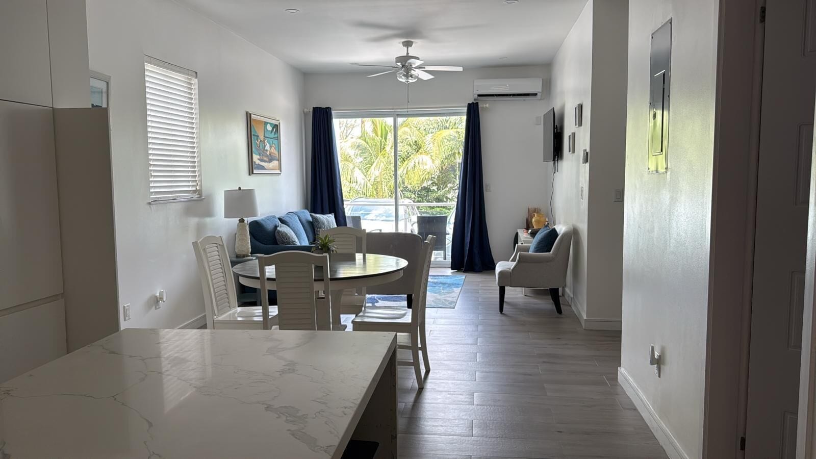 Un espace repas moderne avec une table en marbre, des chaises blanches, un ventilateur de plafond et une vue sur l’extérieur grâce à une grande fenêtre agrémentée de rideaux.