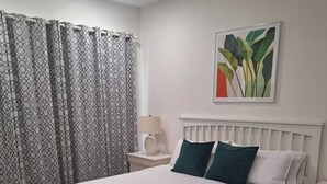 Une chambre à coucher comprenant un lit, une table de chevet, une lampe, une fenêtre avec des rideaux et un tableau accroché au mur.