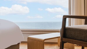Quarto de hotel com uma cama, um banco de madeira e uma grande janela com vista para o mar.