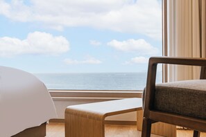 A hotel room with a bed, a wooden bench, and a large window offering a view of the sea.