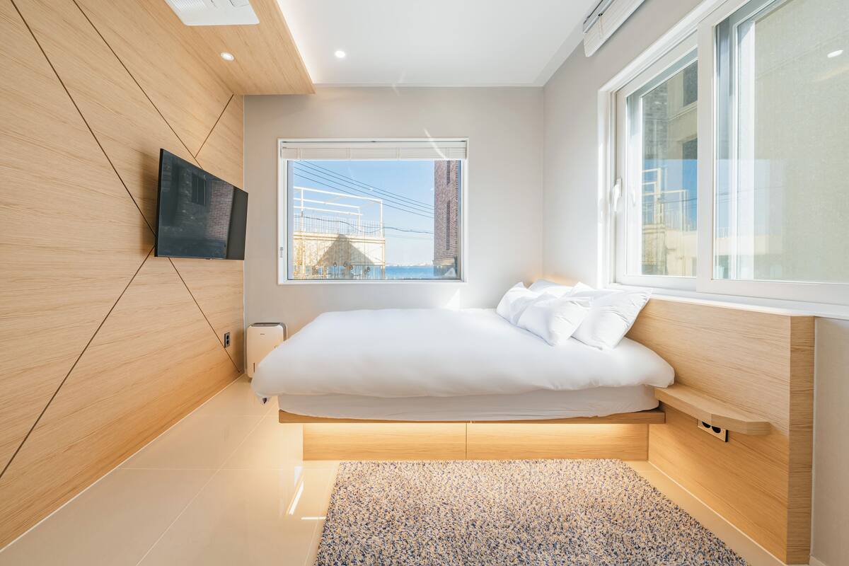 Une chambre moderne avec un lit, un téléviseur fixé au mur et une grande fenêtre.