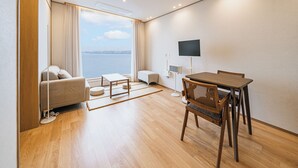 Quarto de hotel moderno com sofá, mesa de jantar, cadeiras, TV e vista para o mar.
