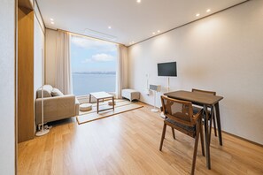 A modern hotel room with a sofa, a dining table, chairs, a TV, and a view of the sea.