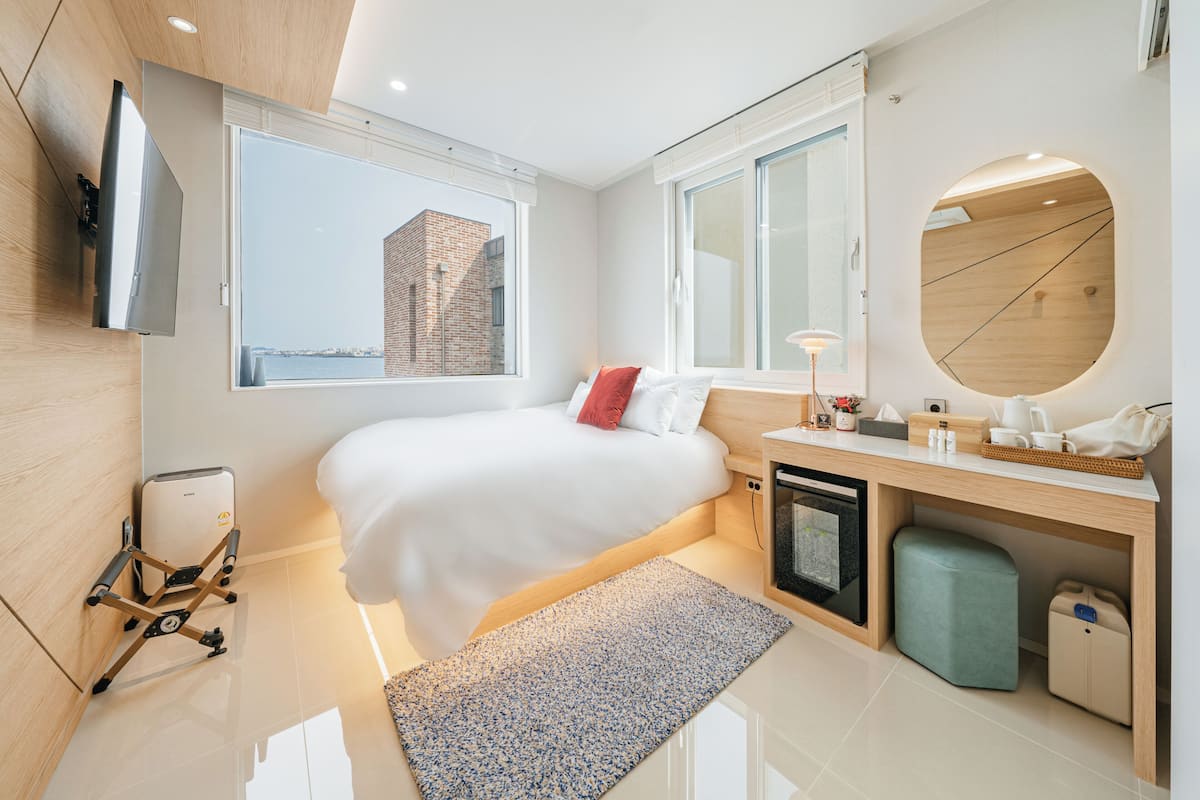 Une chambre d’hôtel moderne équipée d’un lit, d’un bureau, d’un petit réfrigérateur, d’une télévision et d’une grande fenêtre donnant sur la ville.