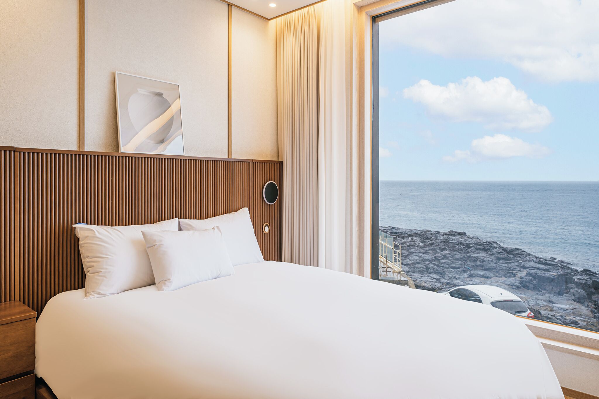 Habitación de hotel con una cama grande, cabecera de madera, mesita de noche, una obra de arte colgada en la pared y vista al océano a través de un ventanal.