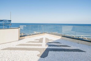 A rooftop terrace with a clear view of the sea, a glass railing, and a pebble-patterned floor.