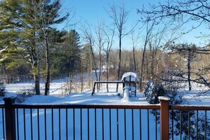 Ein mit Schnee bedeckter Deck mit Holzgeländer, einer Bank und einem ebenfalls verschneiten Gazebo in bewaldeter Landschaft.