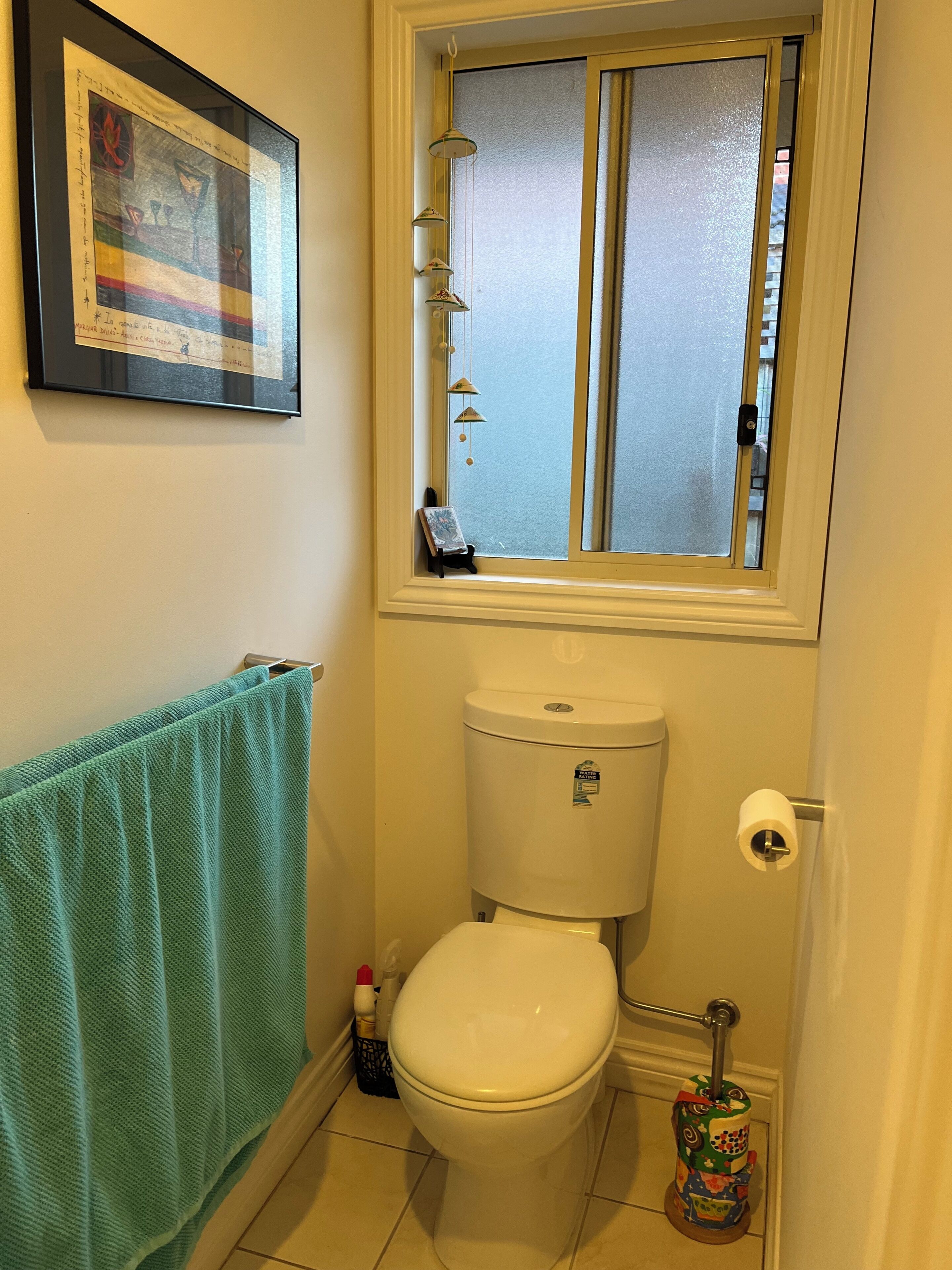 A bathroom with a toilet, a window with a view, a framed picture on the wall, and a towel rack.