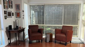 Een ruime woonkamer met houten vloeren, twee gestreepte fauteuils, een bijzettafeltje en een groot raam met jaloezieën.