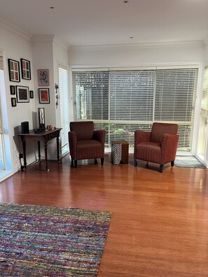 A spacious living room with hardwood flooring, two striped armchairs, a small side table, and a large window with blinds. - Light bright and airy, garden outlook from every window, quiet location (Glen Iris)