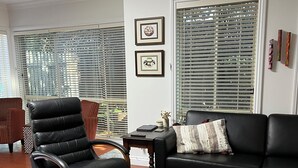 Een woonkamer met een zwarte leren bank, een zwarte lederen fauteuil, een getextureerd vloerkleed, een salontafel met tijdschriften en een raam met jaloezieën.