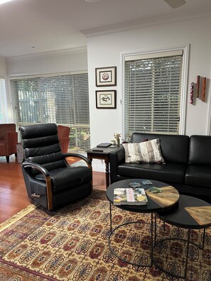 A living room with a black leather sofa, a black leather chair, a patterned rug, a coffee table with magazines, and a window with blinds. - Light bright and airy, garden outlook from every window, quiet location (Glen Iris)